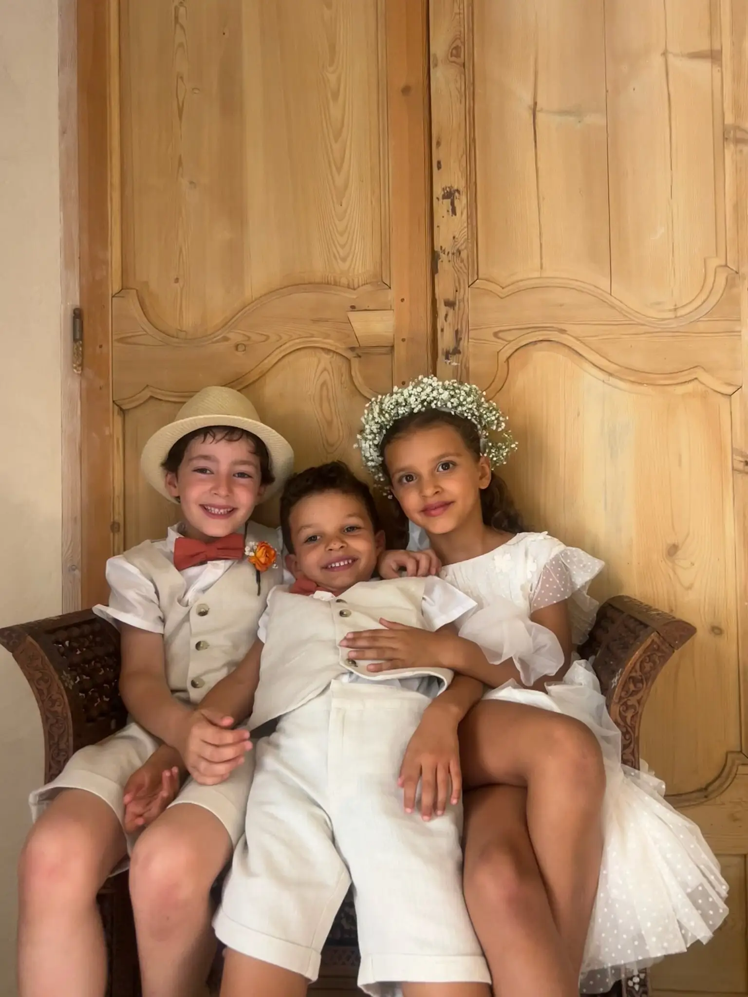 Trois enfants en tenue de fête sourient, assis devant une porte en bois